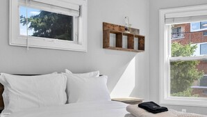 Quarto com cama, roupa de cama branca, janela e prateleira de madeira com um vaso.