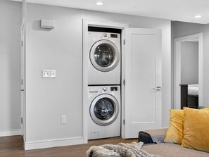 A laundry room with two stacked washing machines, a bed with yellow pillows, and a wooden floor. - Luxe 3 bd | Boston | Penthouse (Boston)