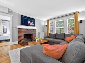 Een woonkamer met een open haard, een grijze zithoek, een salontafel en een flatscreen televisie die boven de haard is bevestigd.