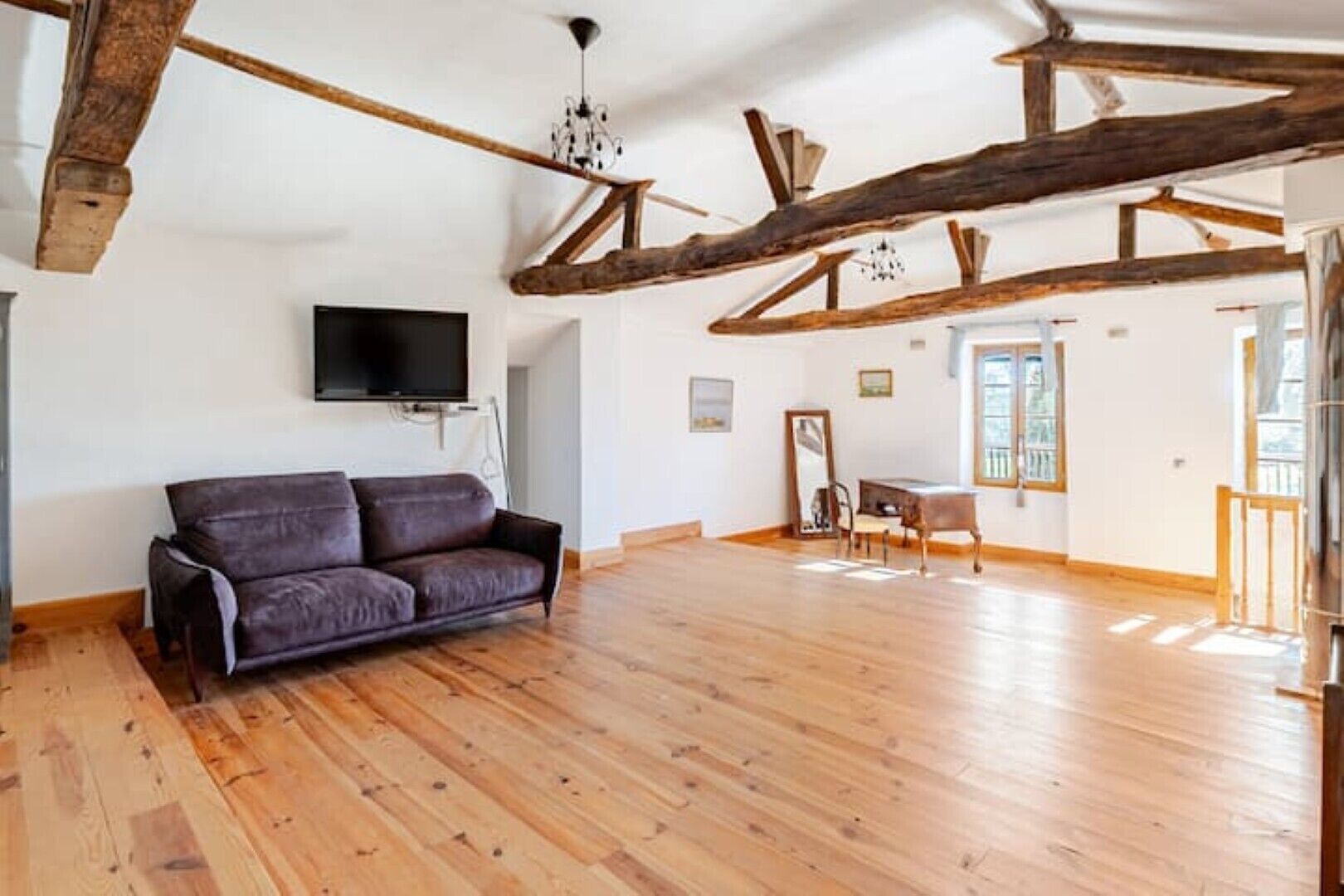 A spacious room with wooden beams, a sofa, a piano, and a TV.