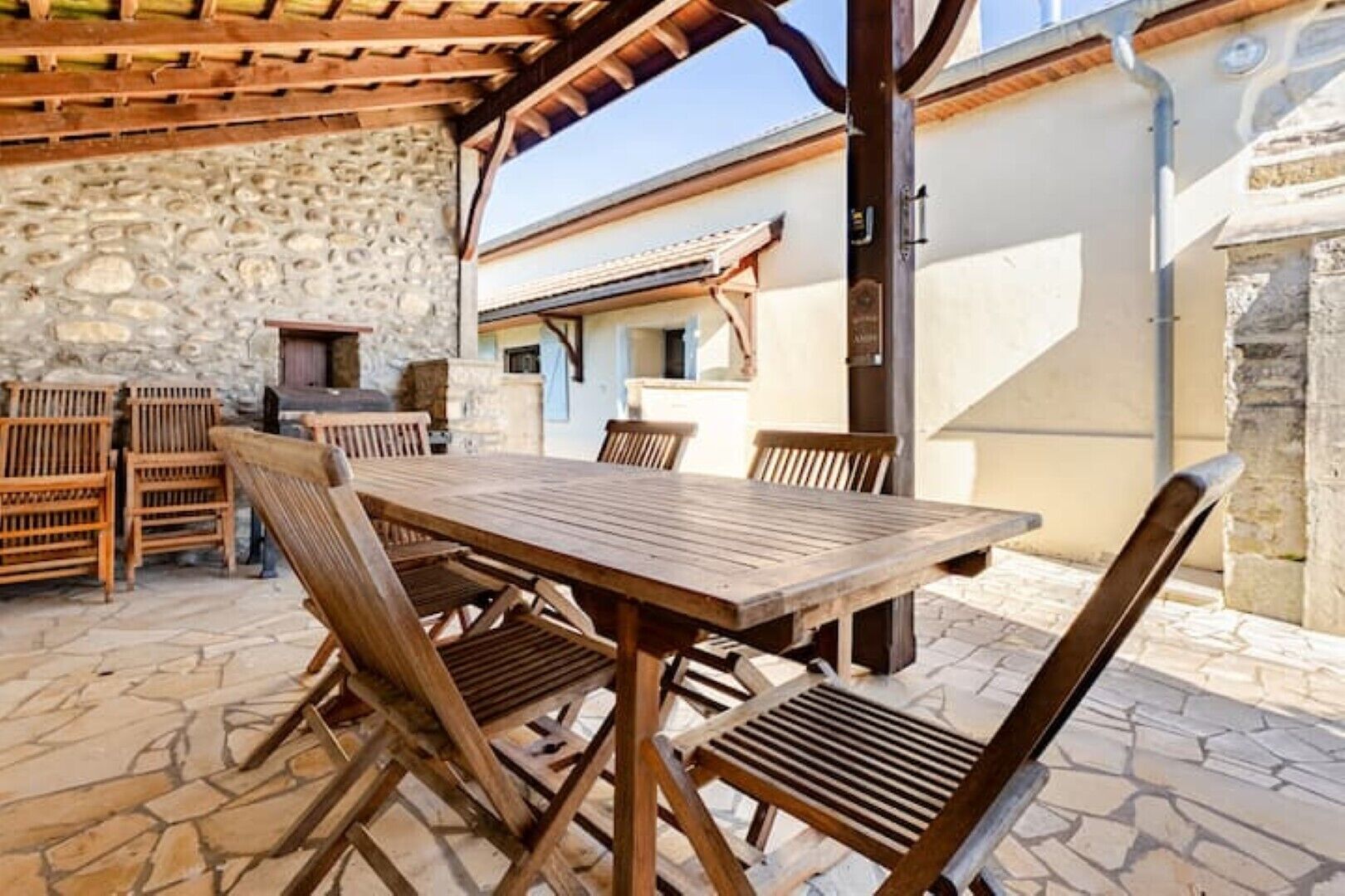 An outdoor patio with a stone floor, wooden table and chairs, and a stone wall.