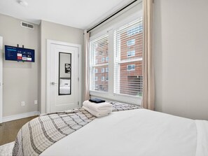 A bedroom with a bed, a TV mounted on the wall, a door, and a window with blinds. - NEW! LUX 3BR w/ 400 Mbps Wifi | ROKU TV & Games! (Boston)