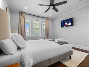 A bedroom with a bed, a ceiling fan, a TV, and a window with curtains.