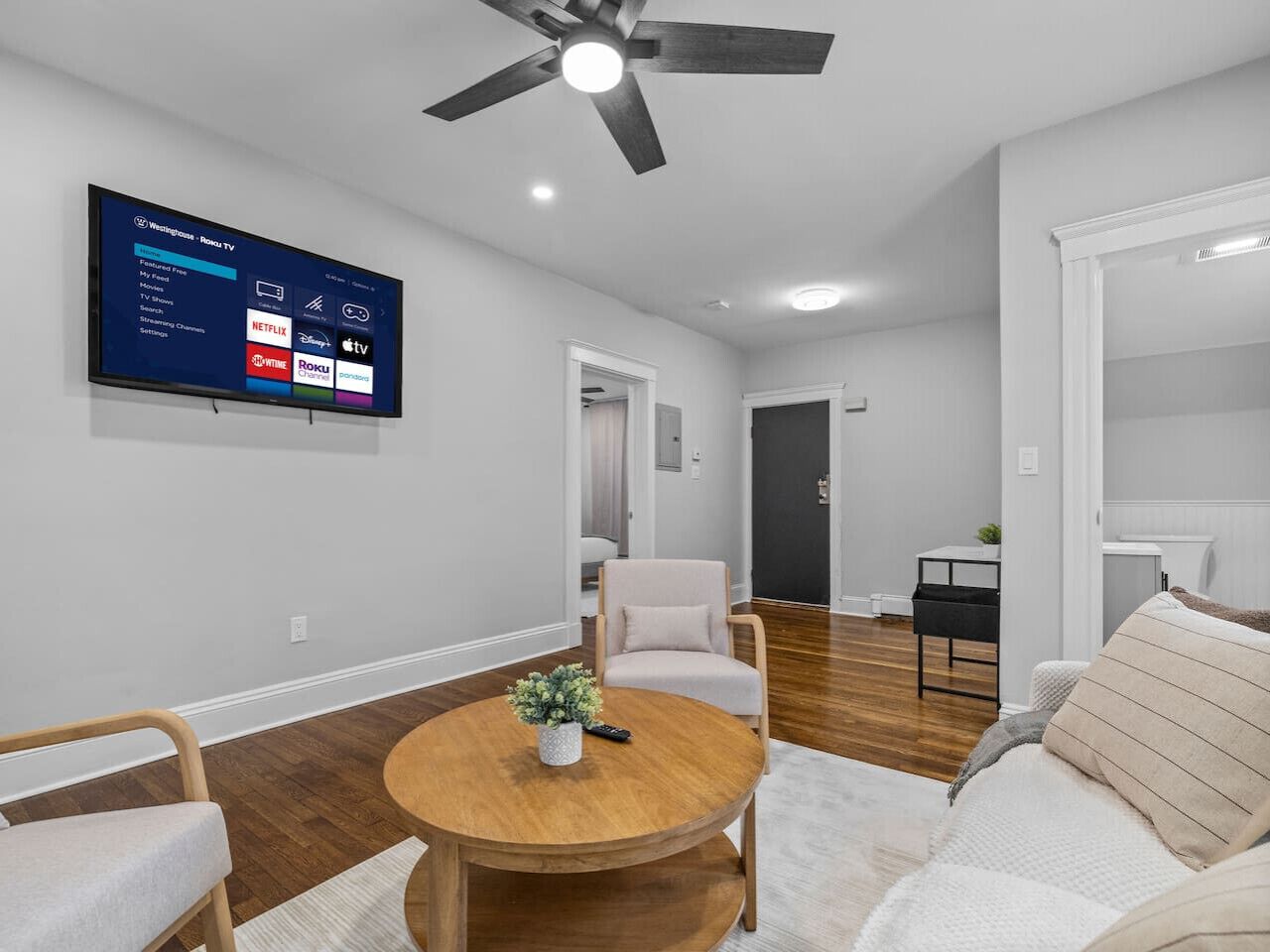 A living room with a round wooden coffee table, a beige sofa, a white armchair, and a television mounted on the wall displaying streaming services.