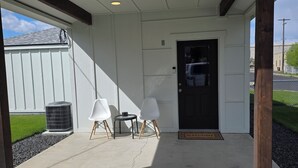 Un espacio al aire libre techado con una puerta, dos sillas blancas, una mesita y un felpudo.