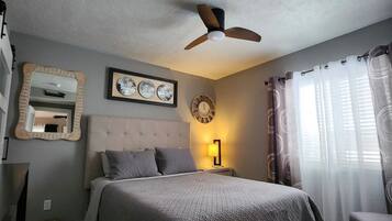 Un dormitorio con cama, ventilador de techo, mesita de noche con lámpara, espejo y ventana con cortinas.