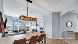 Uma sala de estar moderna com área de refeições, uma janela grande com cortinas, um conjunto de sofás e um espelho redondo na parede.