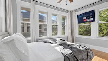 Een slaapkamer met een bed, een plafondventilator, grote ramen en een aan de muur bevestigde televisie waarop streamingdiensten worden weergegeven.