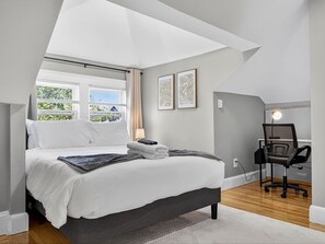 A bedroom with a large bed, a desk, and a chair, with a view of greenery outside.