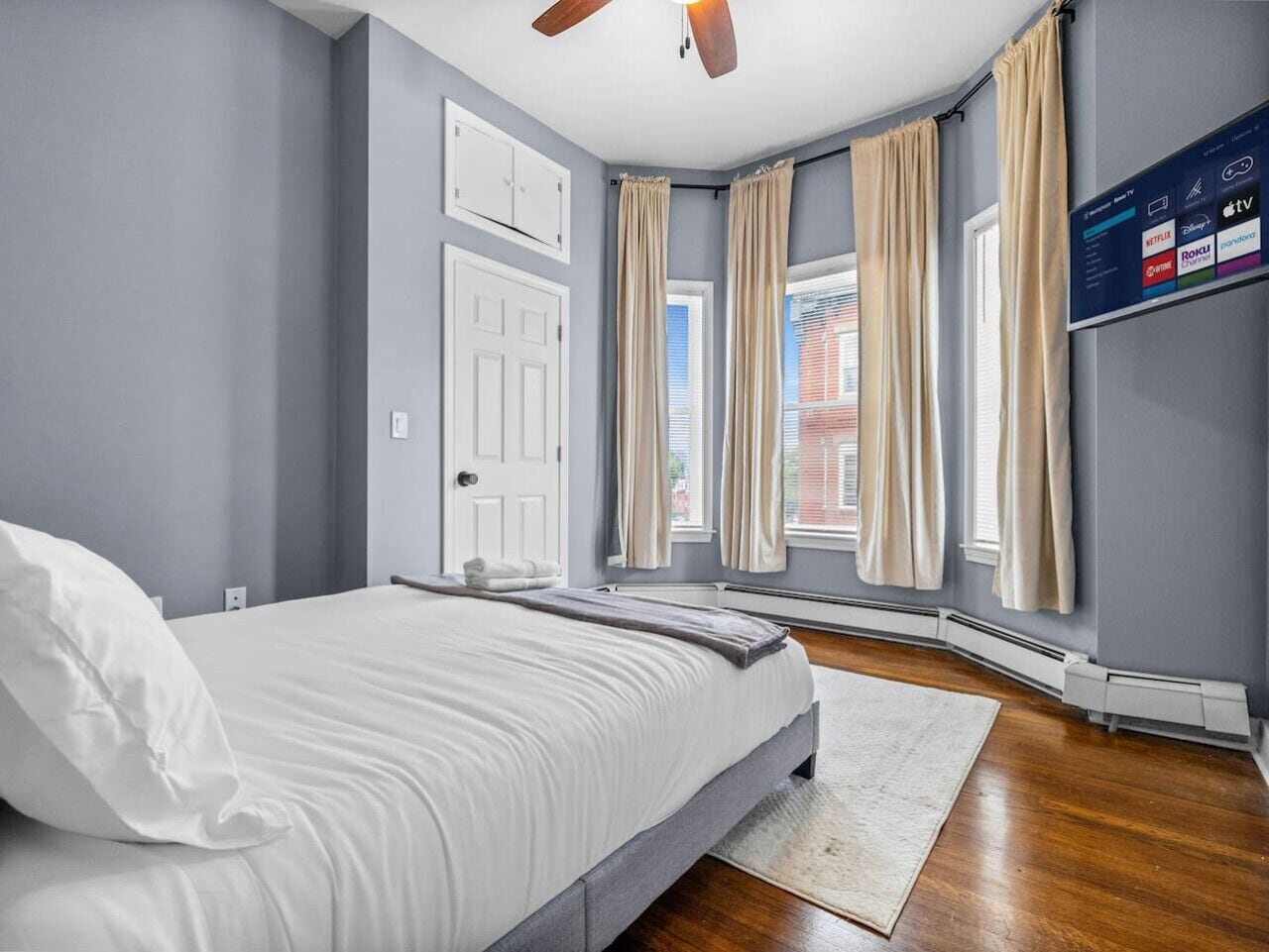 A bedroom with a bed, a ceiling fan, a TV mounted on the wall, and a window with curtains.