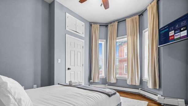 Un dormitorio con cama, ventilador de techo, televisor montado en la pared y ventana con cortinas.