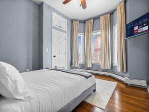 Quarto com cama, ventilador de teto, TV fixada na parede e janela com cortinas.