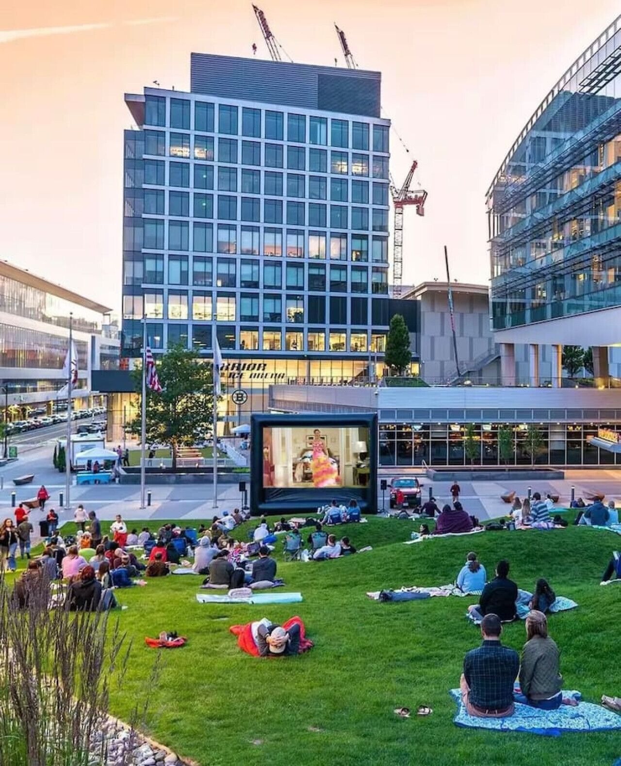Een modern gebouw met een groot scherm waarop een evenement wordt vertoond. Mensen zijn verzameld op een gazon ervoor.