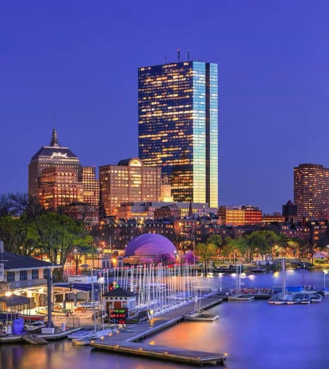 A city skyline at dusk with illuminated buildings, a waterfront promenade, and a dome structure.