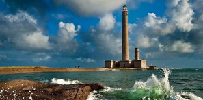 Ein Leuchtturm mit hohem Turm, einem Sockel mit zahlreichen Fenstern und einer felsigen Küste mit brechenden Wellen.