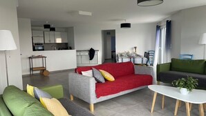 Un salon moderne avec un canapé gris, un plaid rouge et un canapé vert. On aperçoit une table à manger avec des chaises et, au fond, une cuisine.