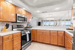 A modern kitchen with wooden cabinets, stainless steel appliances, and a large window offering a view. - Shoreline Towers 1094-seasonal beach service-pool (Destin)
