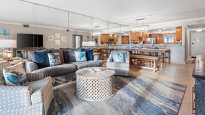 Una sala de estar moderna con sofá, sillón y reposapiés, un comedor con mesa y sillas, y una cocina con gabinetes de madera y electrodomésticos.
