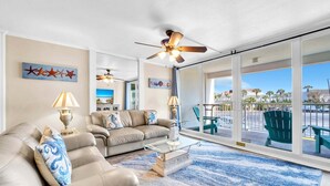 A spacious living room with a large glass door leading to an outdoor patio, a ceiling fan, and a view of the outside. - Shoreline Towers 3023 -seasonal beach service pool-near beach (Destin)