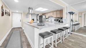 A modern kitchen with a central island, bar stools, and a built-in stove. - Shoreline Towers 3023 -seasonal beach service pool-near beach (Destin)