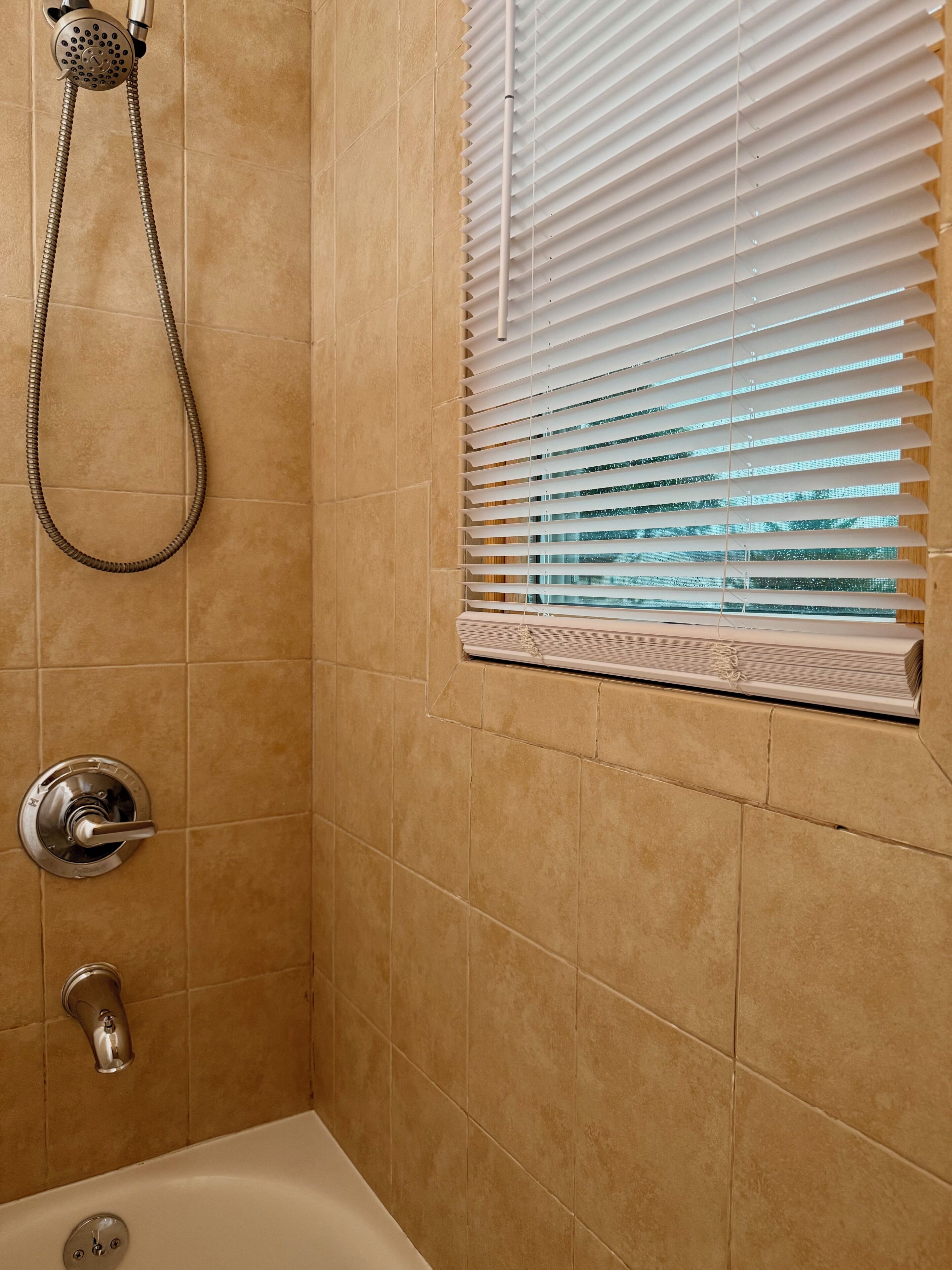 Une salle de bain avec des carreaux de couleur beige, une pomme de douche, une fenêtre avec des stores et une baignoire.