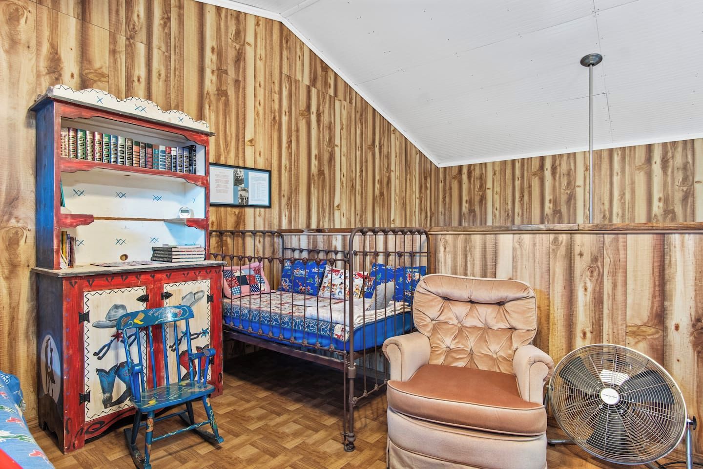 Een kamer met een stapelbed, een schommelstoel, een boekenkast en een ventilator.
