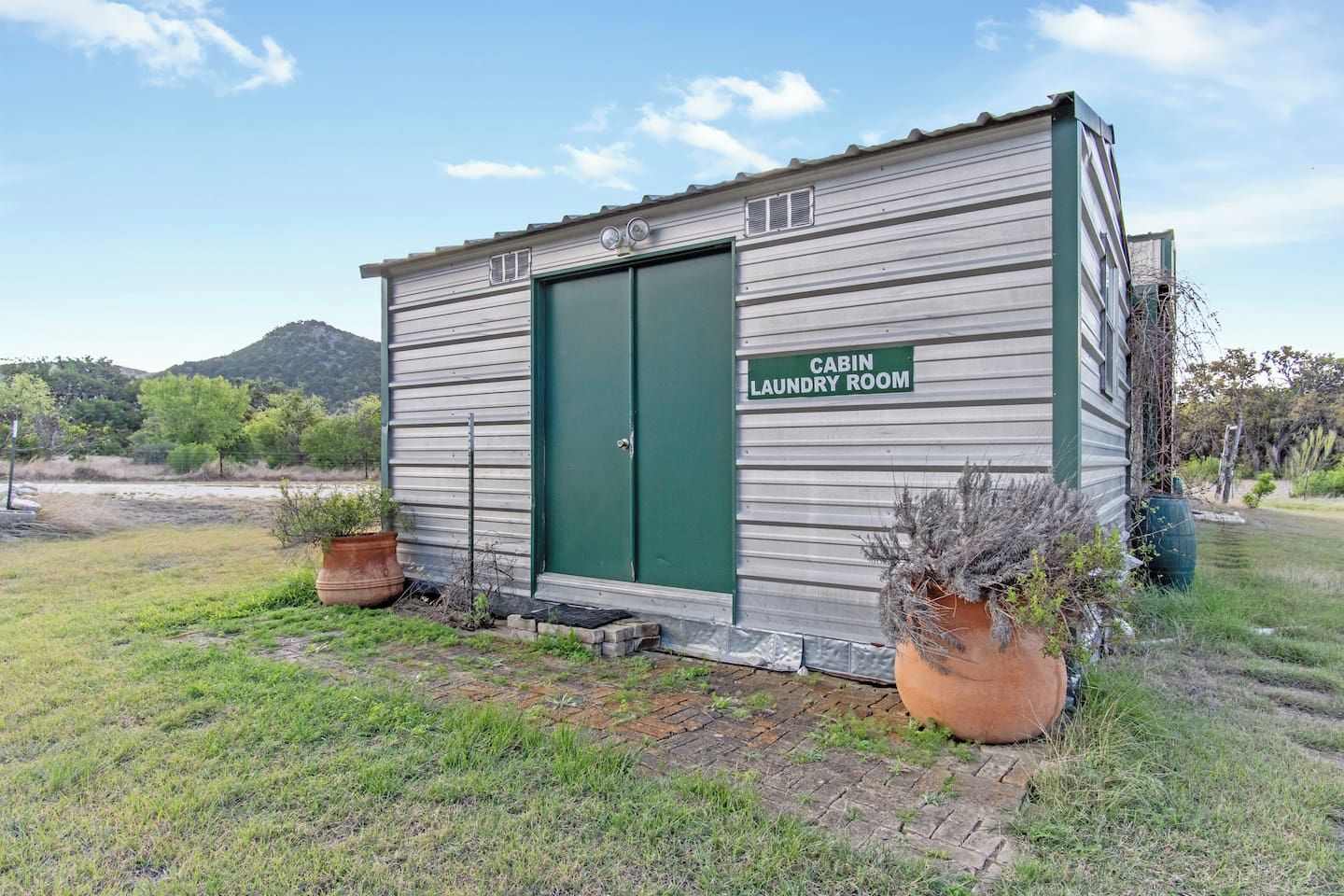 Uma cabana pequena, térrea, com porta verde identificada como 'CABIN LAUNDRY ROOM'. Possui fachada de metal e é cercada por grama e plantas em vasos.