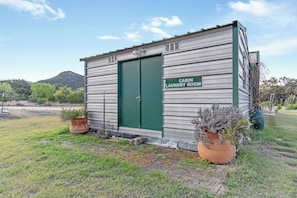 A small, single-story cabin with a green door labeled 'CABIN LAUNDRY ROOM.' It has a metal exterior and is surrounded by grass and potted plants.