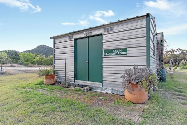 A small, single-story cabin with a green door labeled 'CABIN LAUNDRY ROOM.' It has a metal exterior and is surrounded by grass and potted plants.