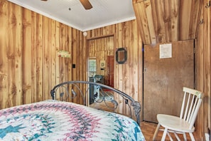 A room with wooden walls, a bed with a floral quilt, a metal-framed bed, a white chair, and a door with a mirror.