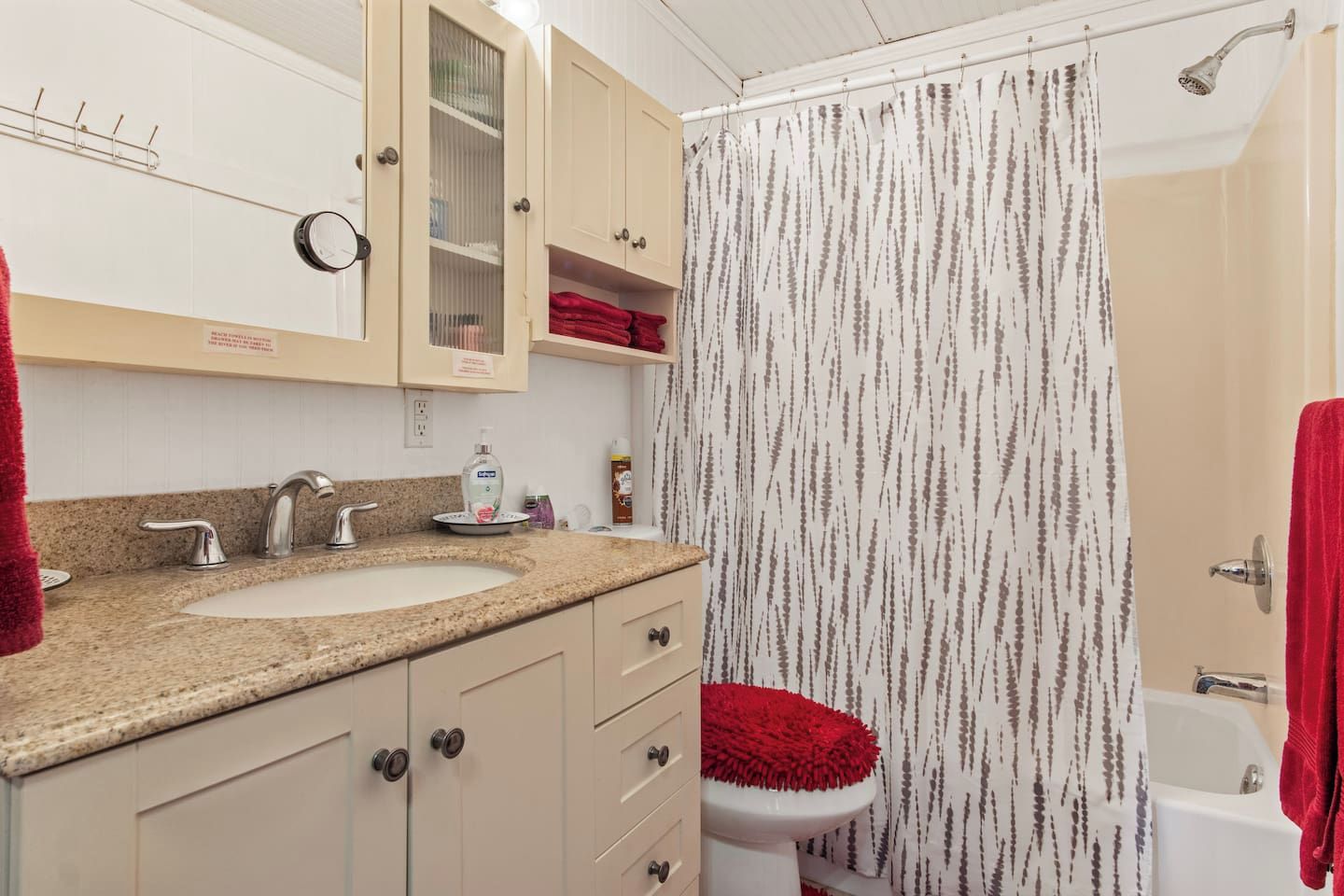 A bathroom with a sink, two faucets, a mirror, a toilet with a red seat, and a shower curtain.