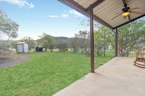 A covered patio with a concrete floor, a ceiling fan, and a view of a grassy yard with trees and a small shed.
