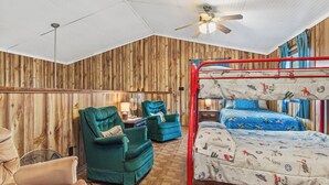 Ein Etagenbett mit rotem Gestell, ein Einzelbett mit blauer Bettdecke, zwei grüne Sessel, eine Holzvertäfelung, ein Deckenventilator und ein Holzboden.