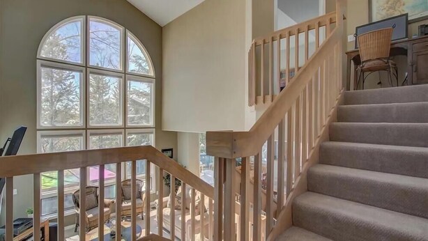 A staircase with wooden railings leading to a second floor, a large arched window with a view of trees, and a chair in the foreground. - Walk To Skiing, Quiet Neighborhood, Big House (Steamboat Springs)