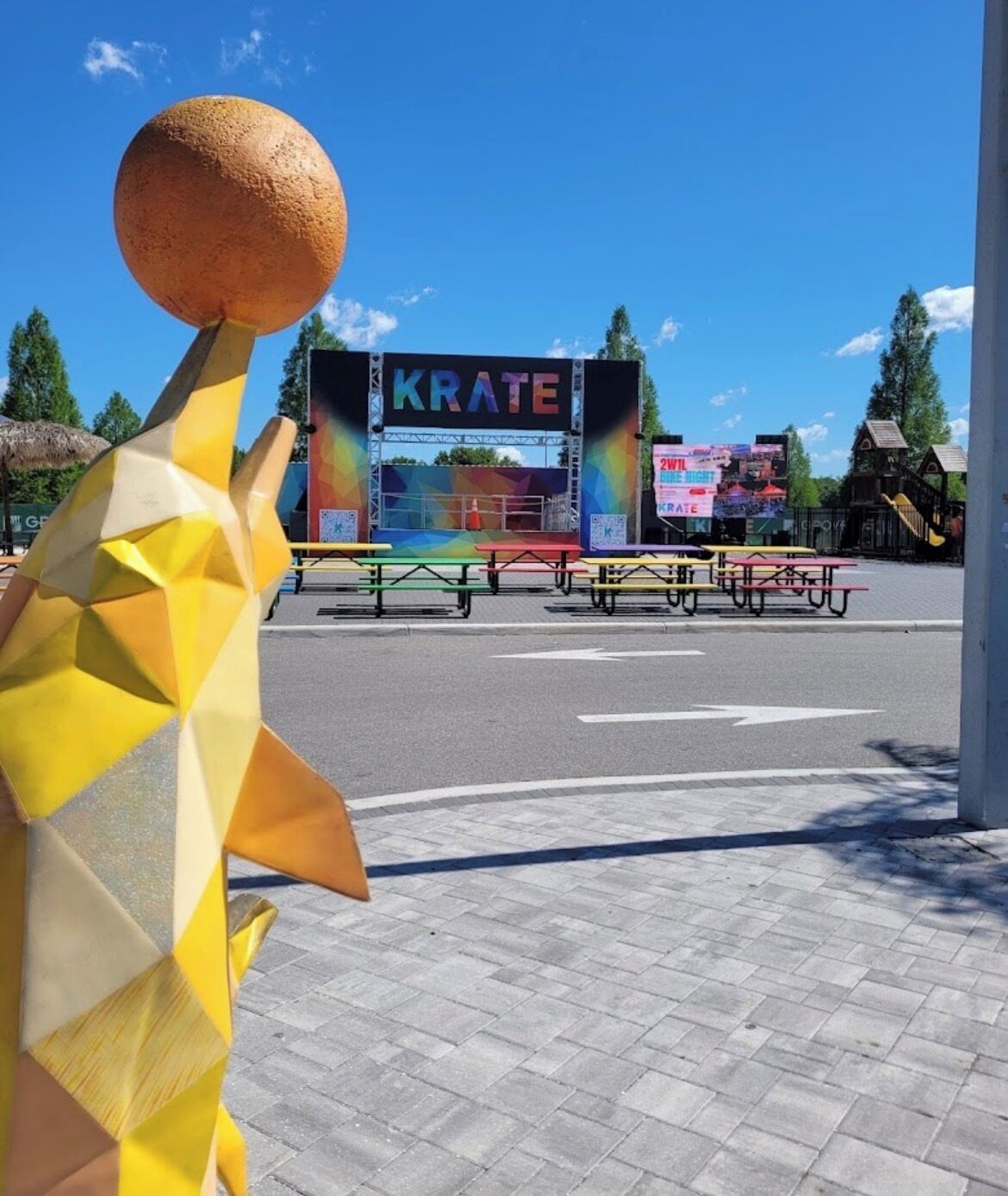 Escultura geométrica em primeiro plano, palco com a palavra 'KRATE' em um painel de fundo, mesas de piquenique e um parquinho ao fundo.