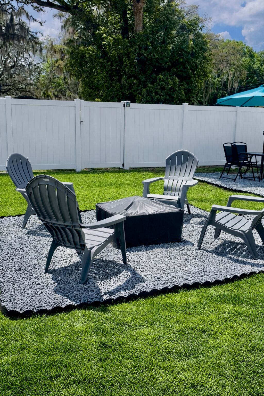 Un área de estar al aire libre con sillas Adirondack y una mesa, rodeada por una valla blanca y follaje.
