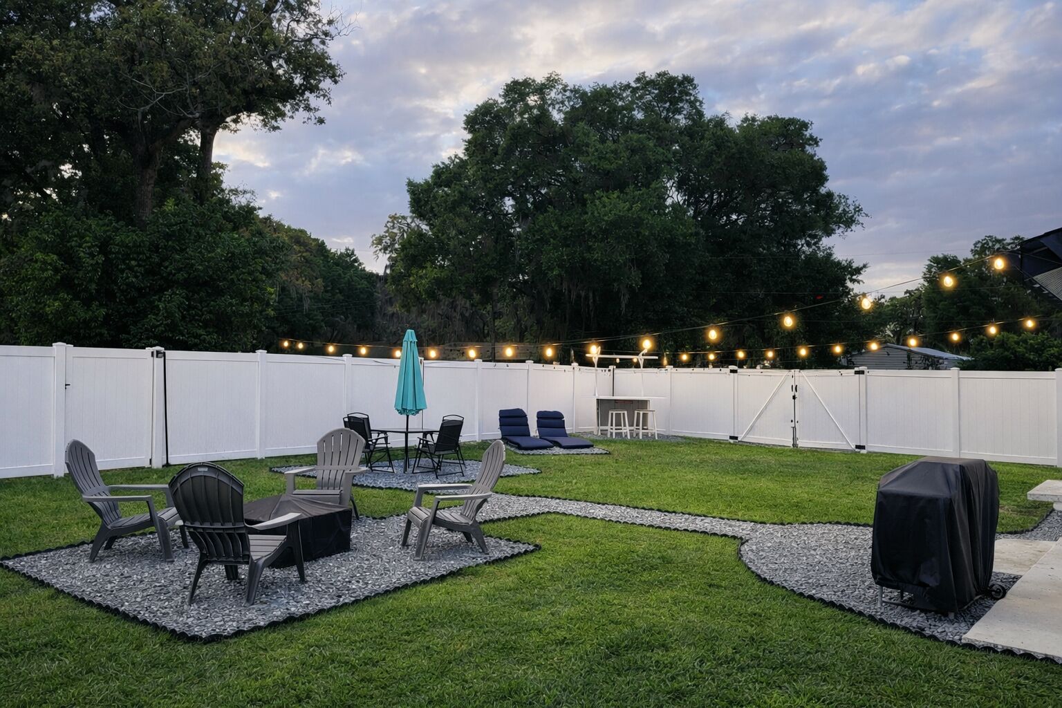A backyard with a white fence, outdoor seating, a grill, and string lights.