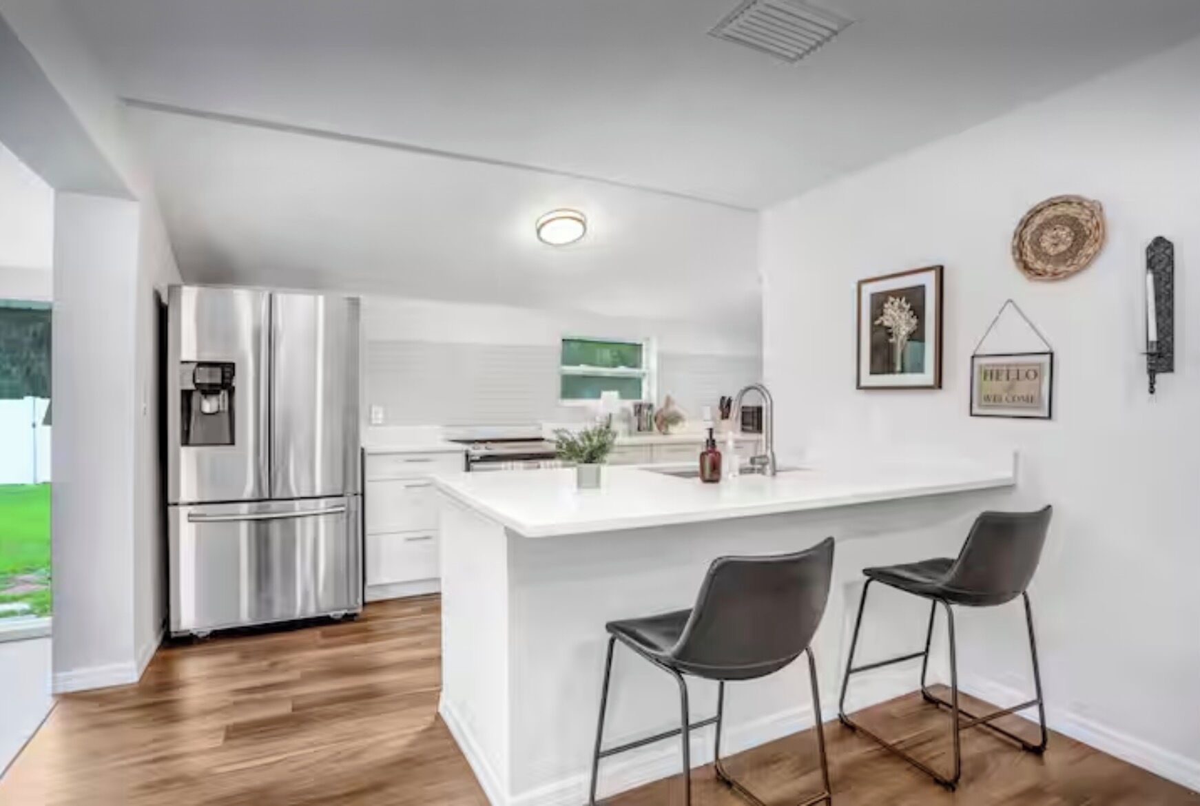 A modern kitchen with a stainless steel refrigerator, a white island with two bar stools, and a view of a green outdoor area.