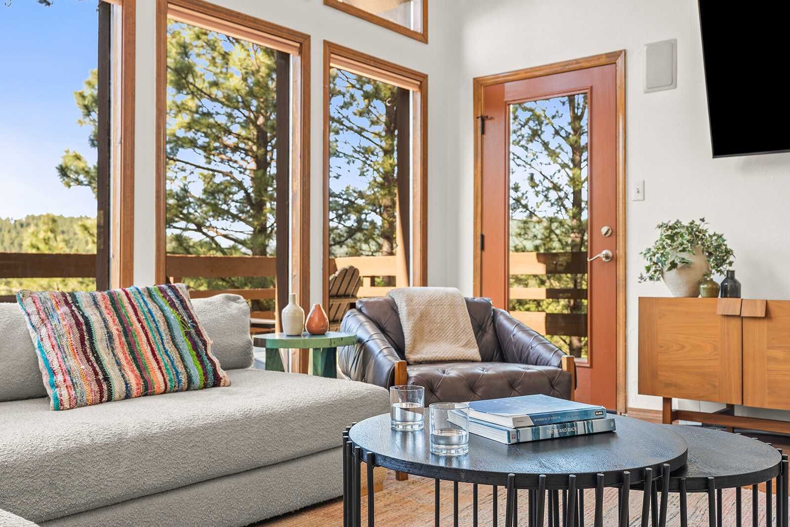 Un salon confortable avec un canapé, un fauteuil et une table basse, de grandes fenêtres donnant sur la verdure et un meuble en bois.