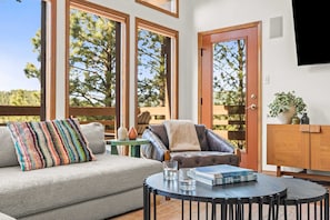 A cozy living area with a sofa, armchair, and coffee table, large windows with a view of trees, and a wooden cabinet. - Mountain-View Cabin | Hot Tub + Cozy Wood Stove (Angel Fire)