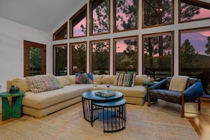 A spacious living room with a large sectional sofa, a round coffee table, and a view of trees through large windows. - Mountain-View Cabin | Hot Tub + Cozy Wood Stove (Angel Fire)