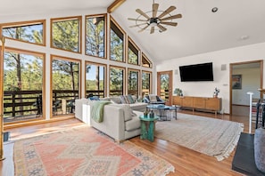 A spacious living room with a large sofa, a coffee table, and a TV. There are large windows and a ceiling fan. - Mountain-View Cabin | Hot Tub + Cozy Wood Stove (Angel Fire)
