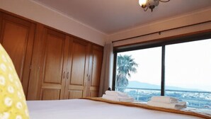 Ein Hotelzimmer mit einem großen Bett, einem Holzschrank und Blick auf Palmen und Berge durch ein großes Fenster.