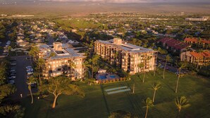 A high-rise hotel with multiple floors, a swimming pool, and a surrounding landscape with trees and a distant mountain range. - 🌅 Maui Magic! Panoramic Beachfront Stay! Ocean Views, Pool, Hot Tub, Sleeps 8🌊 (Kihei)