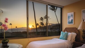 A hotel room with a large window, a bed, a bedside table with a vase, and a wall-mounted lamp. - 🌅 Maui Magic! Panoramic Beachfront Stay! Ocean Views, Pool, Hot Tub, Sleeps 8🌊 (Kihei)