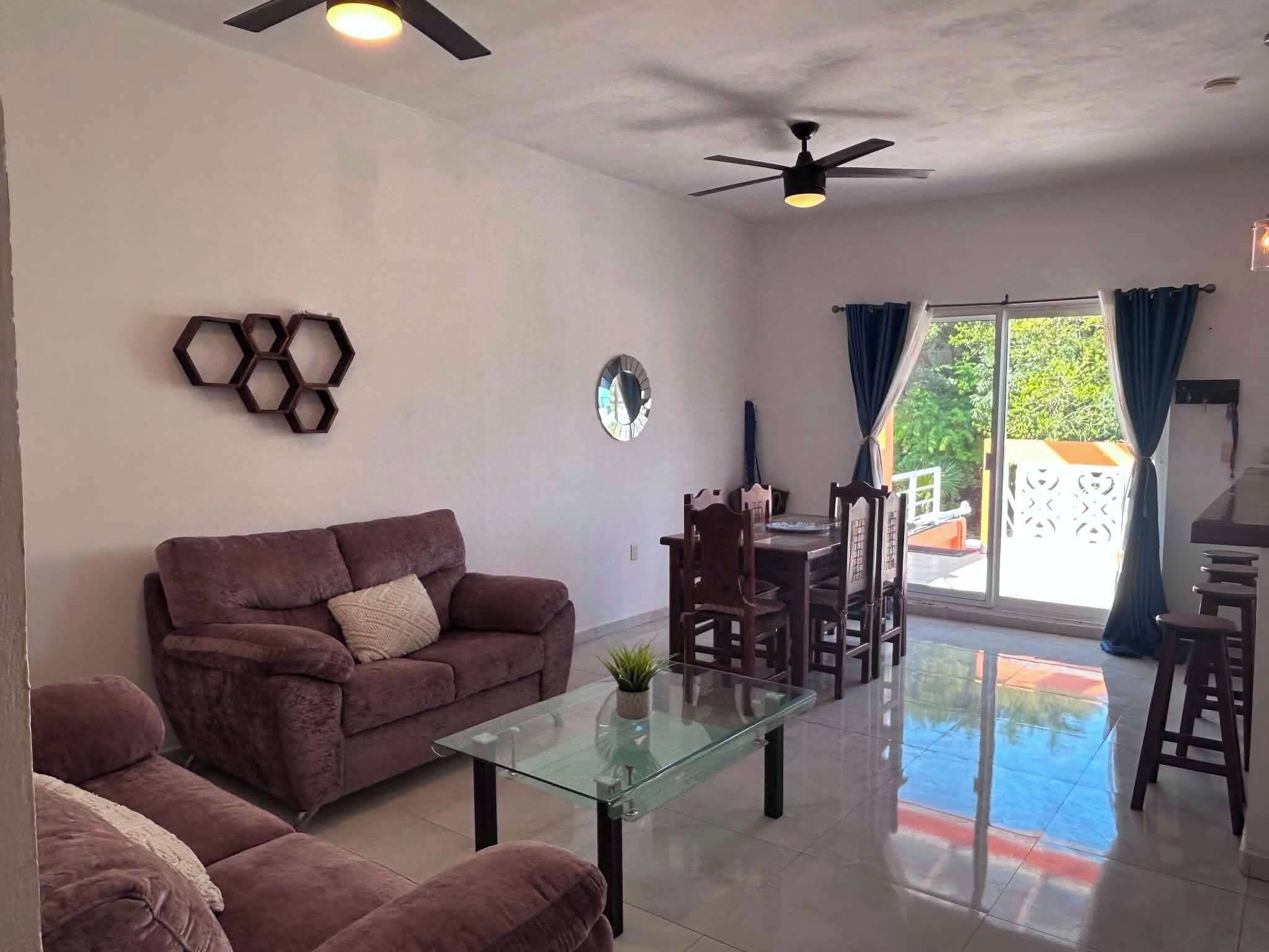 A living room with a sofa, a glass coffee table, a dining area with chairs, and a ceiling fan.