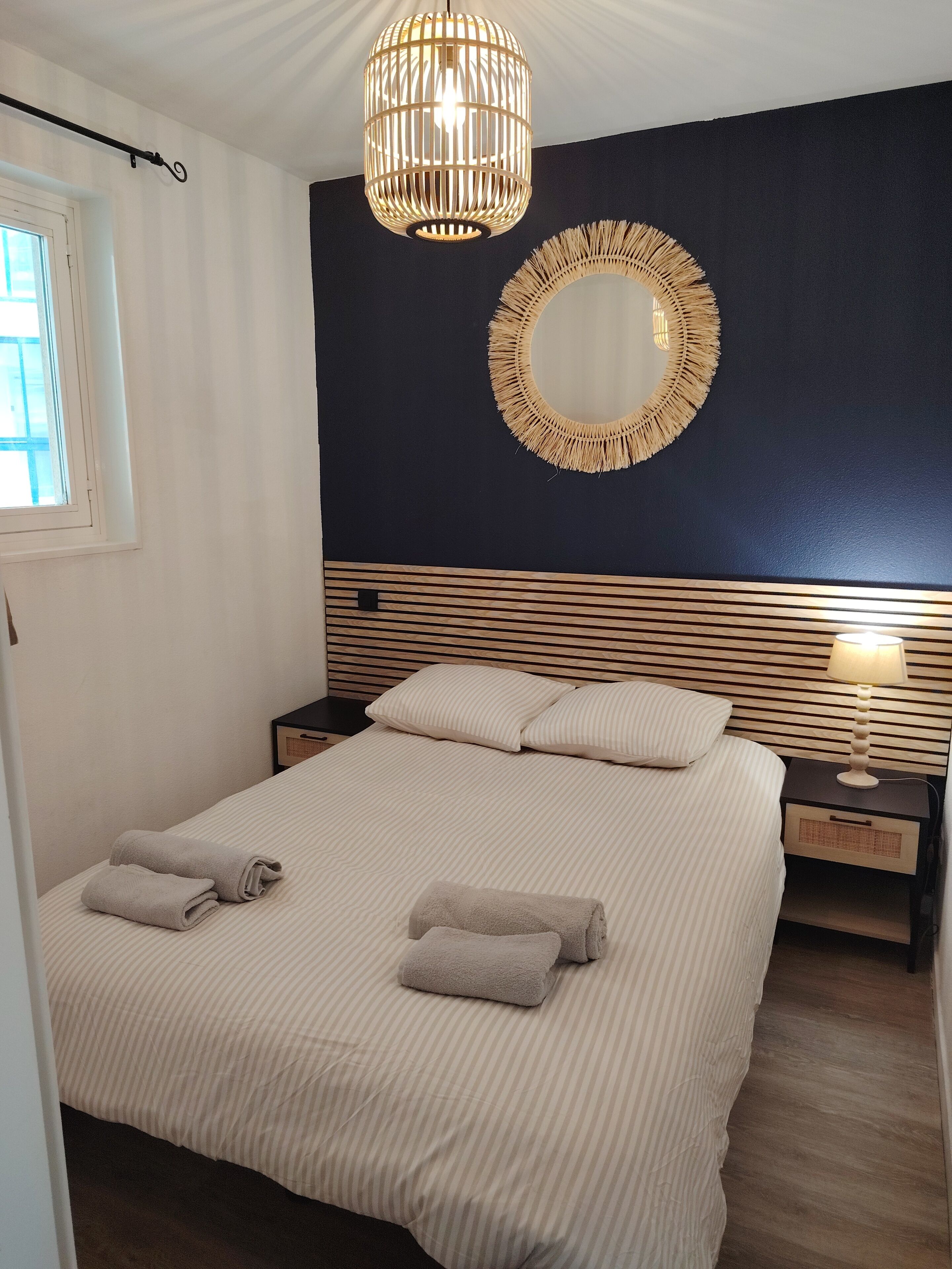 Un dormitorio con cama, dos mesitas de noche, lámpara de noche, lámpara colgante, espejo en la pared y ventana.