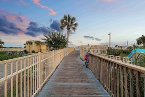 A wooden boardwalk with railings, palm trees, and a distant view of a building.