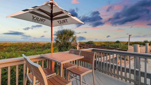 Una terraza de madera con una mesa y sillas, una sombrilla con la palabra “TRUJUY” y vista a la playa y al cielo.