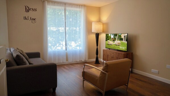 A living room with a sofa, a chair, a TV, a lamp, and a window with sheer curtains. - Sleek & Stylish Downtown LA Studio (Los Angeles)